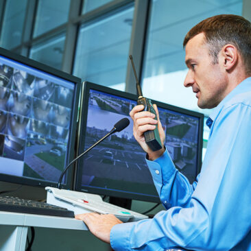Homme à son bureau qui tient un talkie walkie, en train de regarder des images de vidéosurveillance