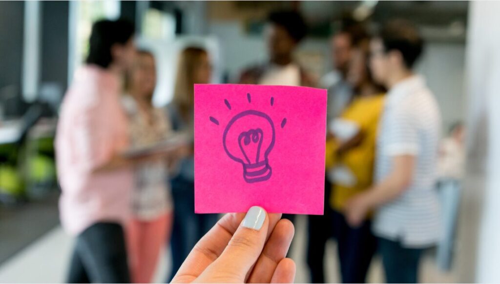 A hand holding a pink post it note with a light bulb drawn on it.
